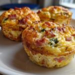 Three golden-brown, freshly baked Egg Muffin Cups with visible pieces of ham and green herbs served on a white plate.