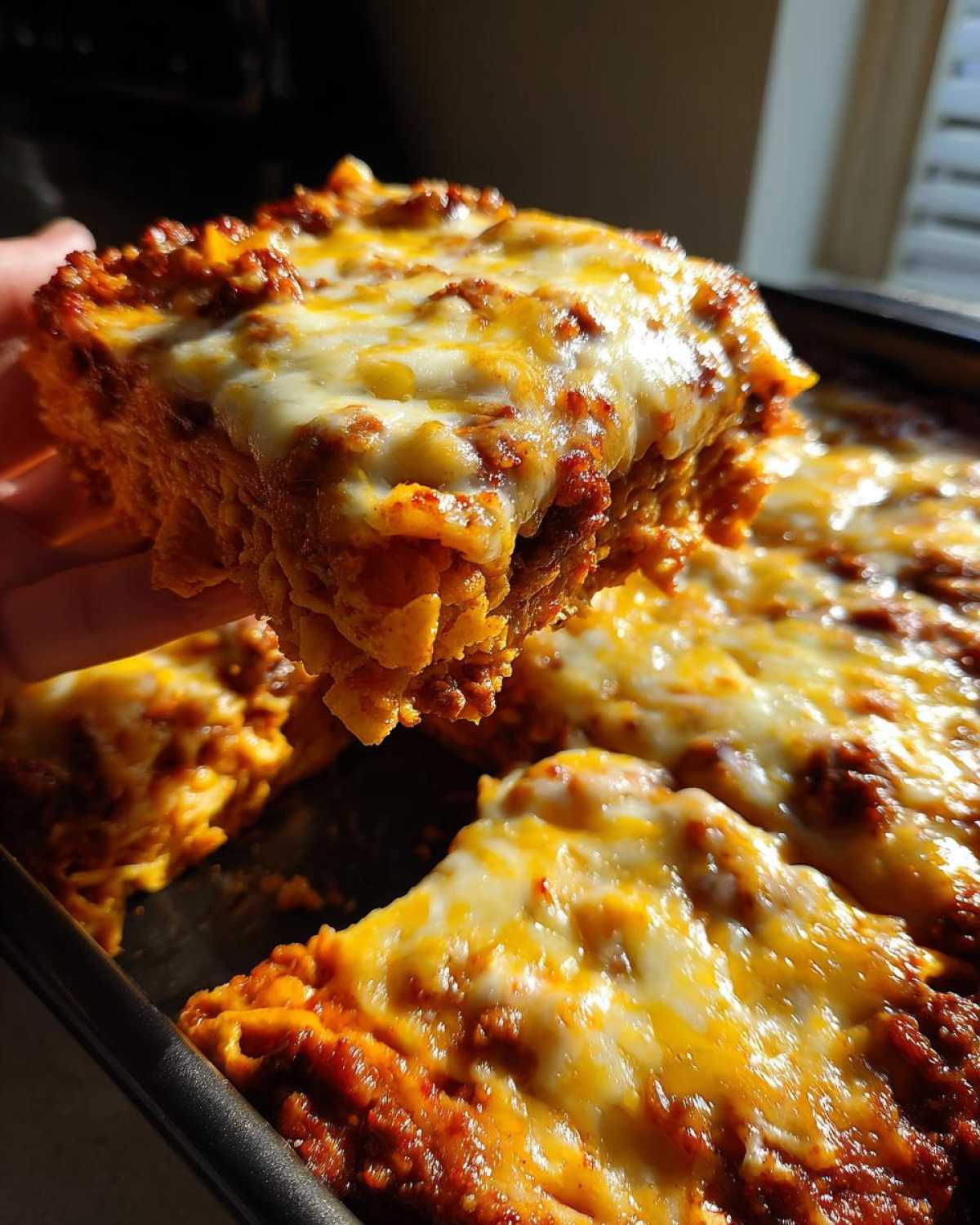 A hand lifts a gooey, cheesy slice of Dorito Casserole topped with melted cheddar and Monterey Jack cheese.