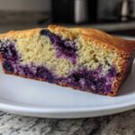 Close-up of a moist slice of blueberry banana bread showing rich purple blueberries baked into the yellow crumb.