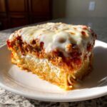 A thick, square slice of baked spaghetti pie with layers of pasta, ricotta, meat sauce, and melted mozzarella cheese on top.