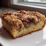 A close-up of a moist slice of sour cream coffee cake featuring a thick cinnamon streusel topping on a white plate.