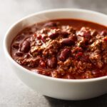 Close-up of a steaming white bowl filled with rich, hearty crockpot chili featuring ground beef and kidney beans.