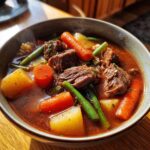 Close-up of a steaming bowl filled with rich vegetable beef soup, featuring chunks of beef, carrots, potatoes, and green beans.