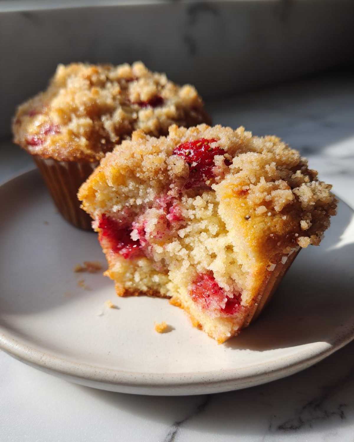 A close-up of a moist strawberry muffin cut in half, revealing bright red fruit and a crumb topping, next to another whole strawberry muffin.
