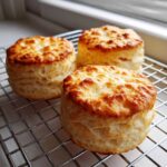 Three golden-brown, tall, fluffy scones cooling on a wire rack near a window.