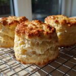 Three tall, fluffy scones with golden-brown, shiny tops cooling on a wire rack in bright sunlight.