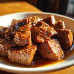 Close-up of tender, saucy cubes of crockpot steak served in a white bowl under warm light.
