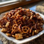 A close-up of a heaping pile of shiny, glazed toffee chex mix recipe ingredients, including pretzels and Chex cereal, served on a white platter.