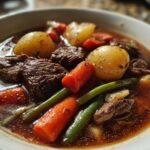 Close-up of a steaming bowl of rich vegetable beef soup featuring tender beef chunks, carrots, green beans, and potatoes.