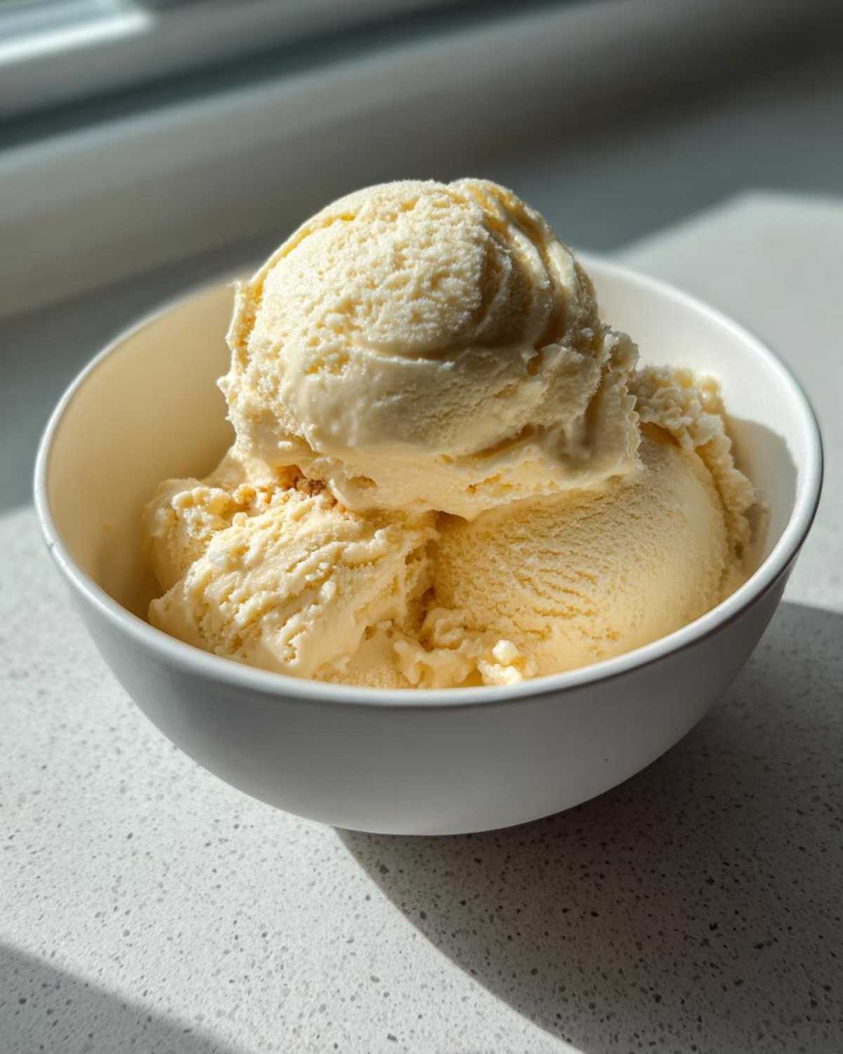 Three scoops of creamy, pale yellow banana ice cream served in a white bowl, brightly lit by sunlight.