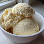Close-up of creamy, pale yellow banana ice cream scooped into a white bowl near a window.