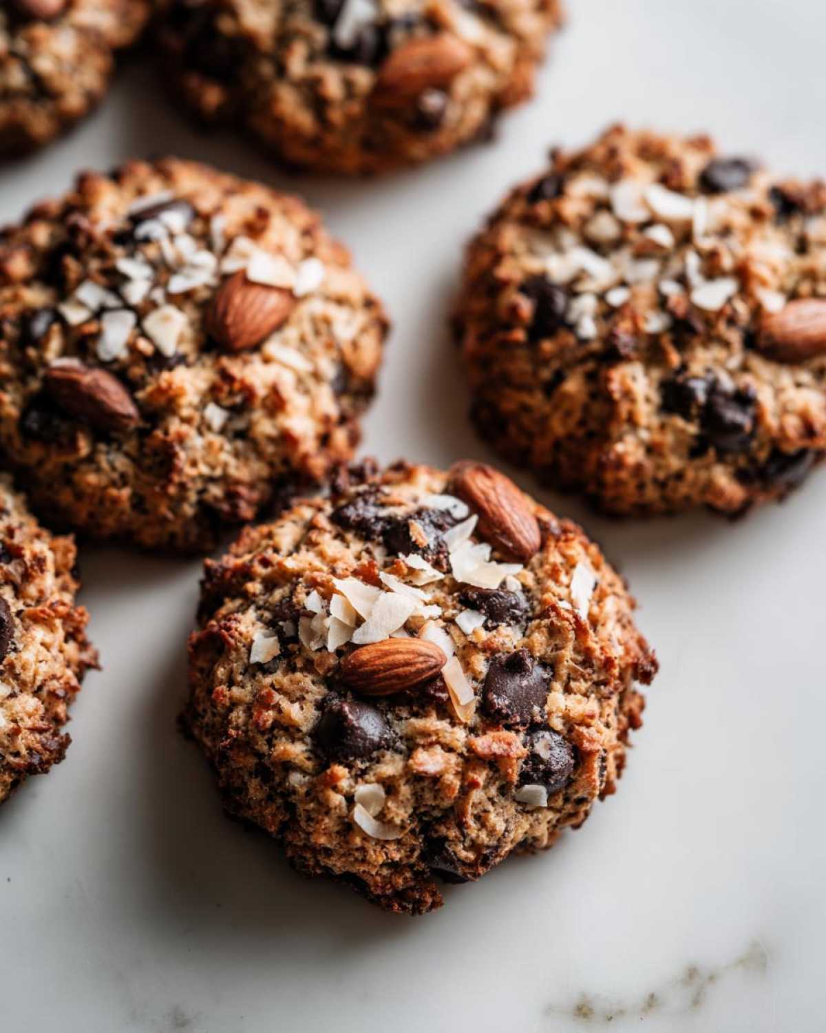 Close-up of freshly baked almond joy cookies topped with whole almonds and coconut flakes on a white marble surface.