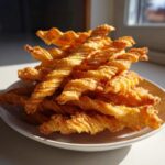 A close-up pile of flaky, golden brown, twisted cheese straws stacked high on a white plate.