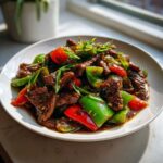 A close-up of a plate of saucy pepper steak featuring strips of beef, bright red and green bell peppers, garnished with scallions.