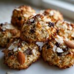 A stack of golden brown almond joy cookies topped with chocolate chips, shredded coconut, and whole almonds.