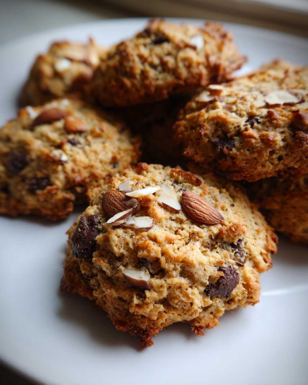 A stack of freshly baked almond joy cookies topped with sliced almonds and visible chocolate chips on a white plate.