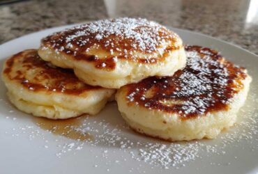 Three fluffy, golden-brown banana pancakes dusted with powdered sugar on a white plate.