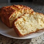 A golden-brown loaf of homemade beer bread, partially sliced, showing the fluffy interior texture.