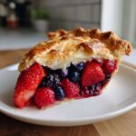 Close-up of a perfect slice of berry pie showing strawberries, raspberries, and blueberries inside a flaky golden crust.