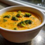 Close-up of a white bowl filled with creamy, orange broccoli cheddar soup topped with bright green broccoli florets.