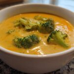 Close-up of a white bowl filled with creamy, orange broccoli cheddar soup topped with bright green florets.