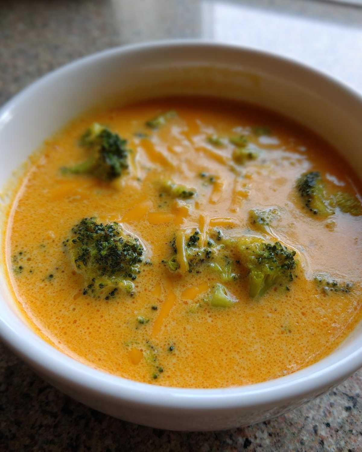 Close-up shot of a white bowl filled with creamy, orange broccoli cheddar soup topped with melted cheese shreds and florets.