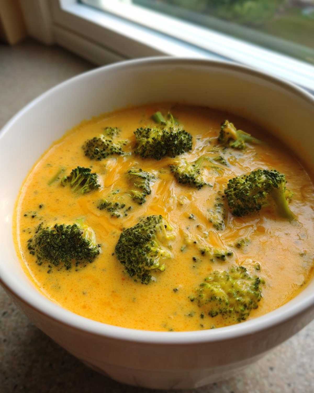 A close-up view of a white bowl filled with creamy, bright orange broccoli cheddar soup topped with fresh broccoli florets.