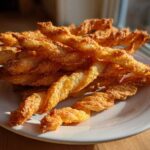 A pile of golden brown, flaky, twisted cheese straws resting on a white plate, catching bright sunlight.