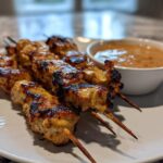 Close-up of grilled chicken satay skewers served on a white plate next to a small bowl of creamy peanut sauce.