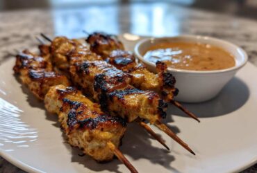 Close-up of grilled chicken satay skewers served on a white plate next to a small bowl of creamy peanut sauce.