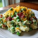 A serving of creamy pea salad mixed with chunks of potato, topped with crispy bacon bits and shredded cheddar cheese.