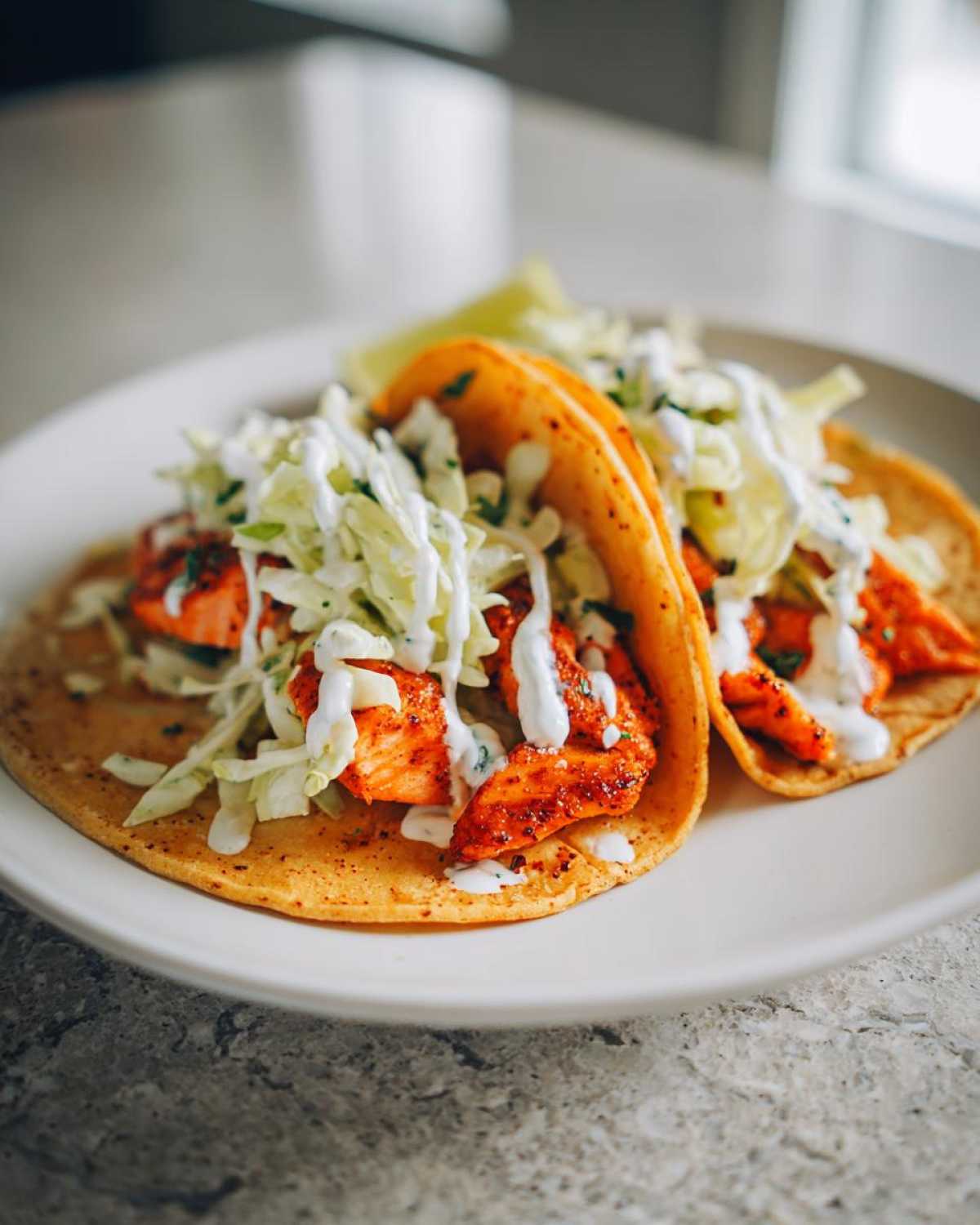 Close-up of two soft tortilla salmon tacos topped with shredded cabbage slaw and drizzled with white sauce.