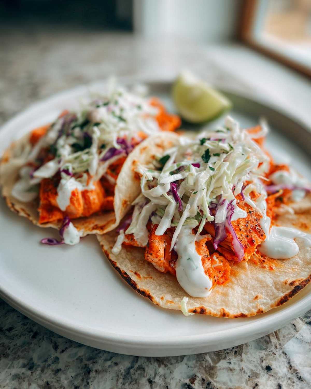 Two appetizing salmon tacos filled with seasoned salmon, topped with creamy sauce and colorful slaw, served with a lime wedge.