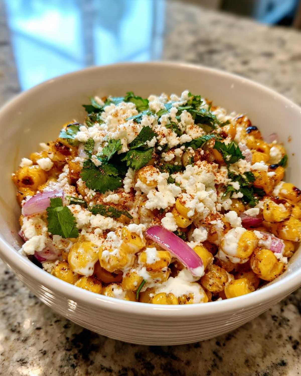 A close-up view of a white bowl filled with amazing street corn salad, topped with cotija cheese, cilantro, and chili powder.