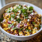 Close-up of a white bowl filled with charred corn kernels, creamy sauce, red onion, cotija cheese, and cilantro, showcasing the street corn salad.