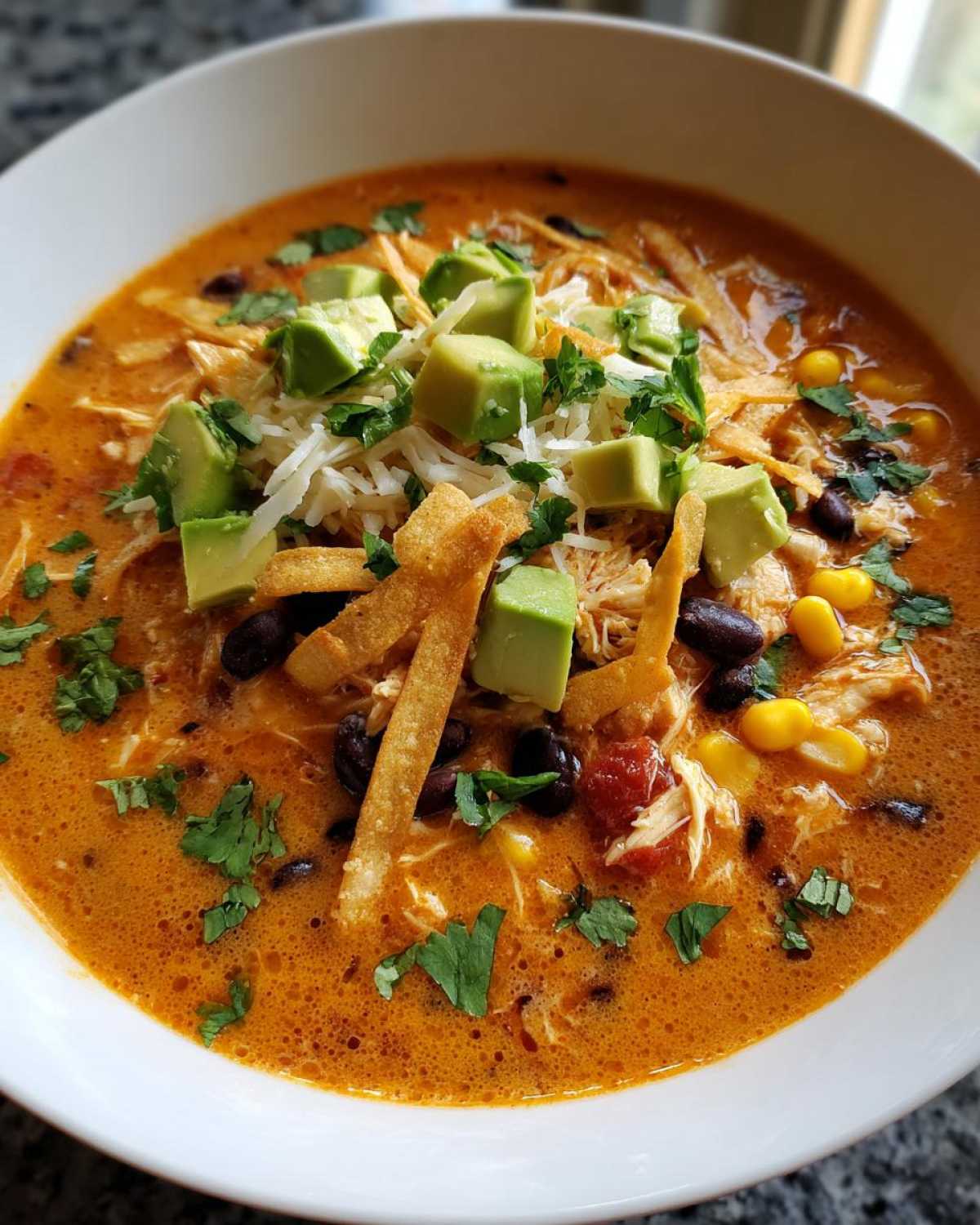 A close-up of a white bowl filled with creamy, orange-hued tortilla soup recipe, topped with avocado, cheese, and tortilla strips.