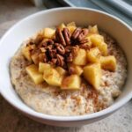A white bowl filled with creamy apple cinnamon oatmeal, topped with diced apples, pecans, and a dusting of cinnamon.