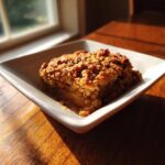 A square serving of warm apple crisp recipe with a golden, crunchy oat topping, served in a white dish.