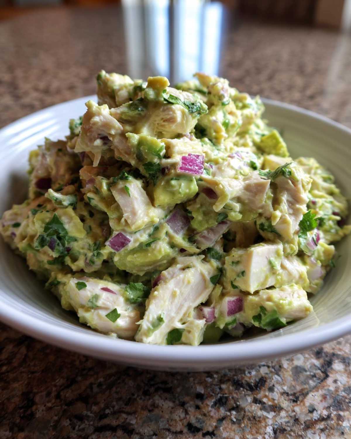 Close-up of chunky avocado chicken salad mixed with red onion and cilantro in a white bowl.