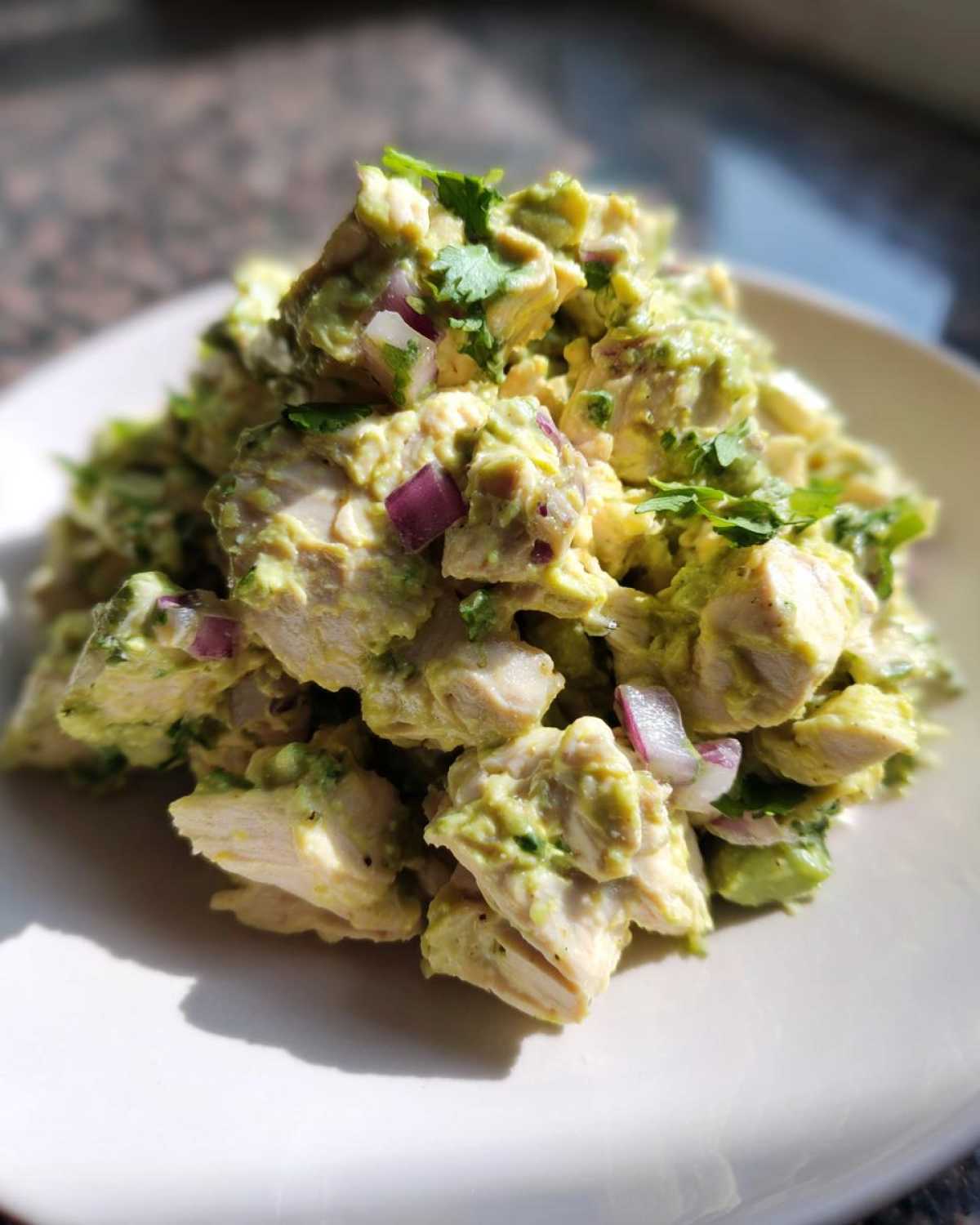 A mound of creamy avocado chicken salad with chunks of chicken, red onion, and cilantro on a white plate.