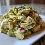A mound of creamy avocado chicken salad featuring chunks of white meat chicken, red onion, and cilantro, served on a white rectangular plate.