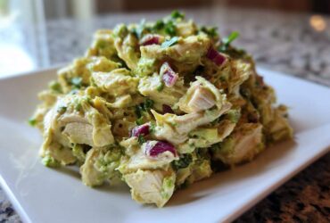 A mound of creamy avocado chicken salad featuring chunks of white meat chicken, red onion, and cilantro, served on a white rectangular plate.