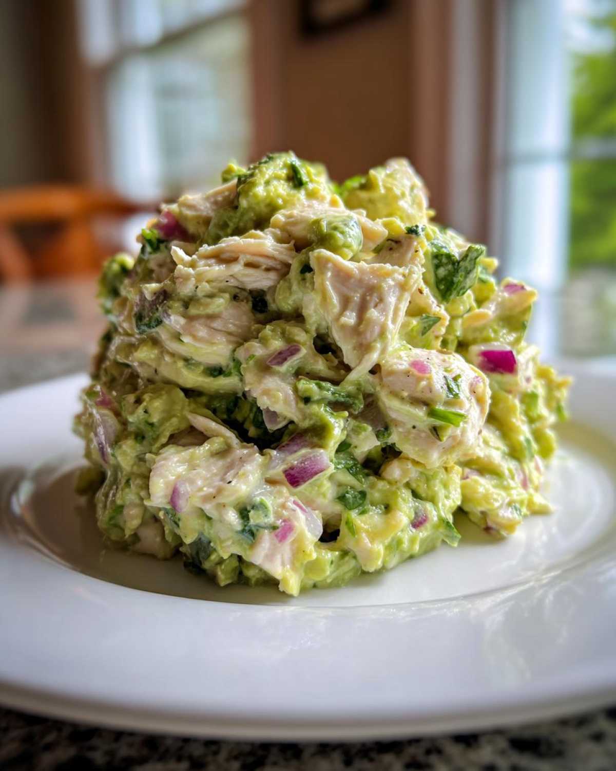 A close-up of a generous scoop of creamy avocado chicken salad mixed with red onion, served on a white plate.