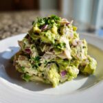 A mound of fresh avocado chicken salad featuring chunks of chicken, diced avocado, red onion, and cilantro, served on a white plate.