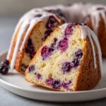 Close-up of a slice of moist breakfast cake filled with dark berries and topped with white glaze.