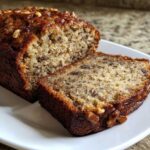 A close-up of the best banana bread, showing a moist crumb and walnuts on top, with one slice cut.