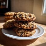 A stack of four golden brown oatmeal cookies, featuring visible oats and raisins, resting on a white plate.