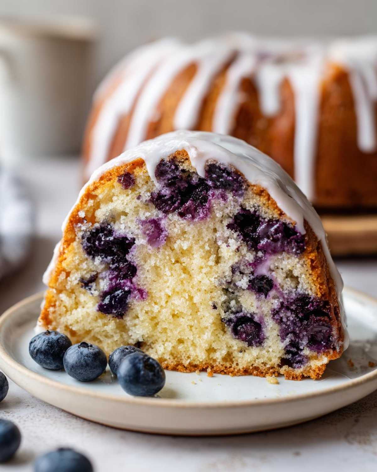 Close-up of a slice of moist blueberry breakfast cake topped with white glaze, served with fresh blueberries.