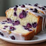 Close-up of a sliced blueberry breakfast cake drizzled with white glaze on a white plate.