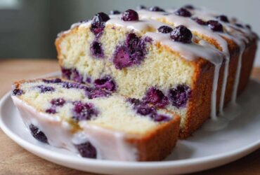 Close-up of a sliced blueberry breakfast cake drizzled with white glaze on a white plate.
